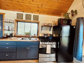 Een kleine keuken met een fornuis, oven en koelkast, houten kastjes en een raam met zonwering.