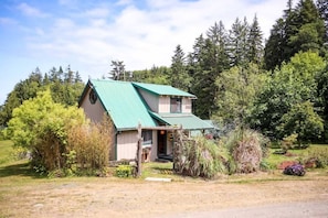 Una pequeña casa de un solo piso con techo verde, rodeada de un bosque denso.
