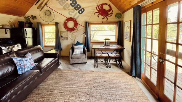 A cozy cabin interior with a wooden ceiling, a dining area, a living room with a leather couch, and a large window with curtains.