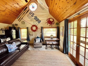 A cozy cabin interior with a wooden ceiling, a dining area, a living room with a leather couch, and a large window with curtains.