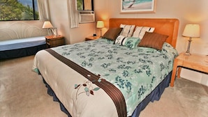 A bedroom with a large bed, a window with a view of trees, a painting on the wall, and two bedside tables with lamps. - Ocean views at Kealia Resort on Sugar Beach! (Kihei)