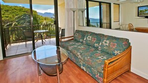 A room with a sofa, a glass table, a balcony with chairs, and a view of greenery and mountains. - Ocean views at Kealia Resort on Sugar Beach! (Kihei)