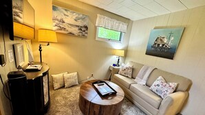 Una sala de estar acogedora con un sofá beige, una mesa de centro de madera y una lámpara. Hay dos cuadros en la pared y una ventana con un encaje blanco.