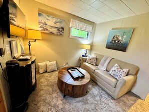 Un salon chaleureux avec un canapé beige, une table basse en bois et une lampe. Deux tableaux ornent le mur, et une fenêtre est agrémentée d’un lambrequin blanc.