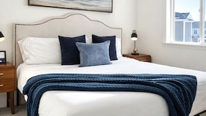 Un lit bien fait, avec une tête de lit blanche, un plaid bleu et plusieurs coussins. Un tableau encadré représentant un paysage est accroché au-dessus du lit.