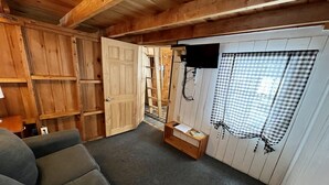 Ein gemütliches Zimmer mit Holzbalkendecke, einem großen Fenster mit karierten Vorhängen, einem an der Wand befestigten Flachbildfernseher und einem kleinen Holztisch mit einem Buch und einer Fernbedienung.