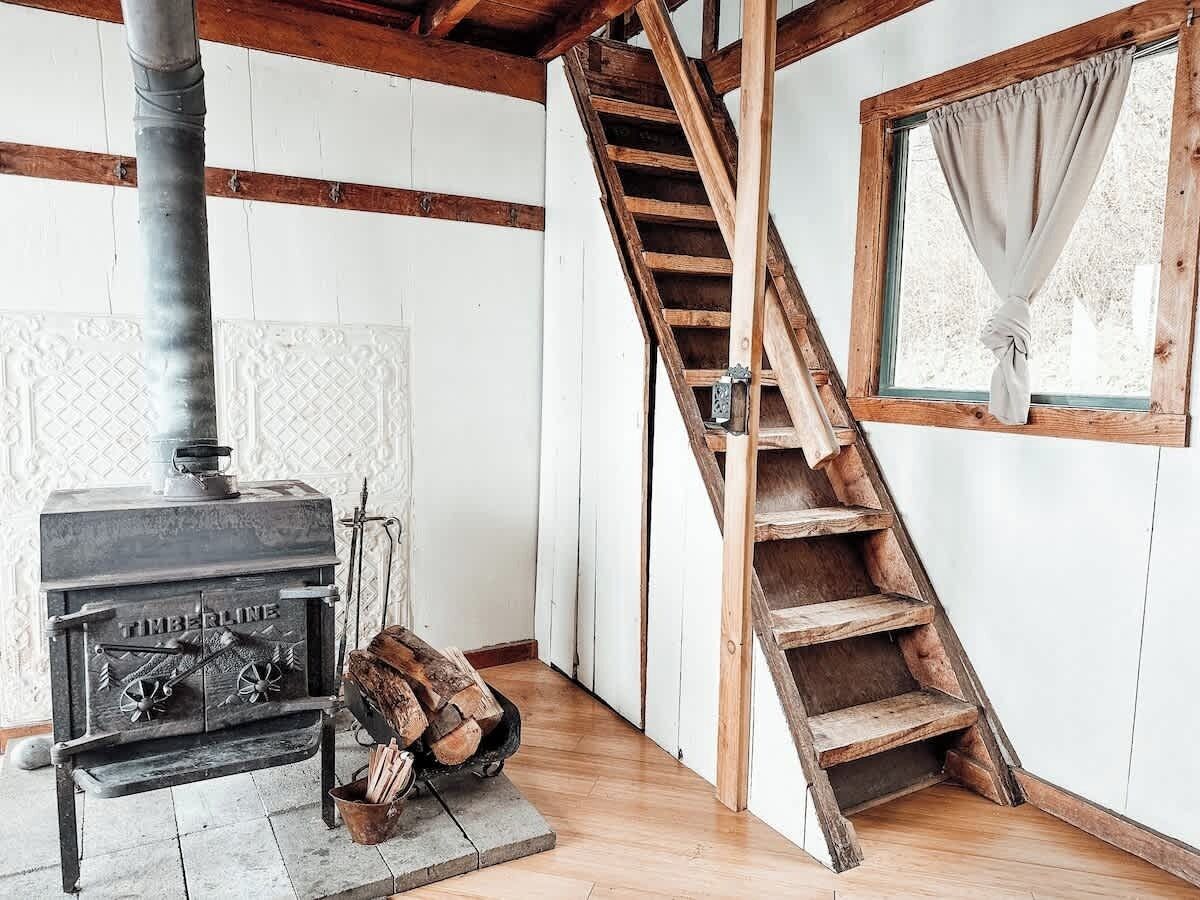 A room with a wood-burning stove, a wooden staircase, and a window with curtains.