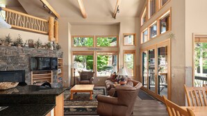 A spacious living room with a stone fireplace, comfortable seating, large windows, and a wooden ceiling. - Gorgeous Townhome Views of the Slopes, Sleeps 12, Private Baths, Private Hot Tub (Whitefish)