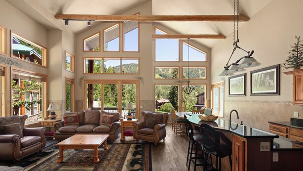 A spacious living area with a large window, a dining area, and a kitchen with wooden beams and a black countertop. - Gorgeous Townhome Views of the Slopes, Sleeps 12, Private Baths, Private Hot Tub (Whitefish)