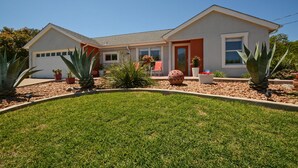 Ett tvåvåningshus med ett garage, en prydlig gräsmatta framför huset med olika växter och en klar, blå himmel.
