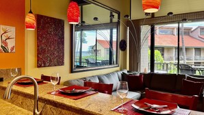 A modern kitchen with granite countertops, a built-in sink, and red accents. There is a dining area with a sofa and chairs, and a large window offering a view of the outside.