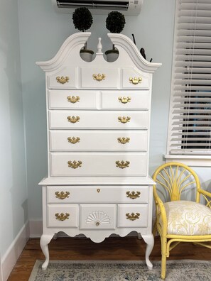 A white dresser with gold handles, a yellow chair with a patterned cushion, and a window with blinds.