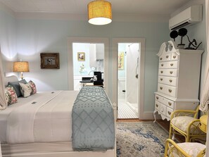 A bedroom with a bed, bedside table, lamp, dresser, chair, and a view into the bathroom.