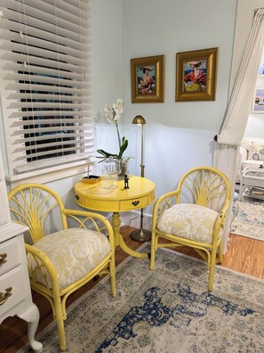 A room with a yellow table, two matching chairs, a white dresser, and a window with blinds. There are two framed pictures on the wall.