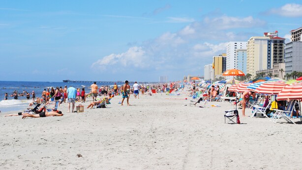 Ein belebter Strand mit Menschen, die sich sonnen und spazieren gehen, Sonnenschirmen und Liegestühlen.