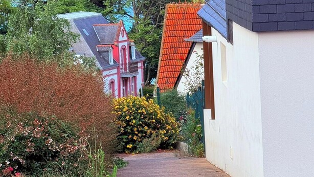A coastal residential area with a pathway leading to a house with a red roof, surrounded by greenery and overlooking the sea.