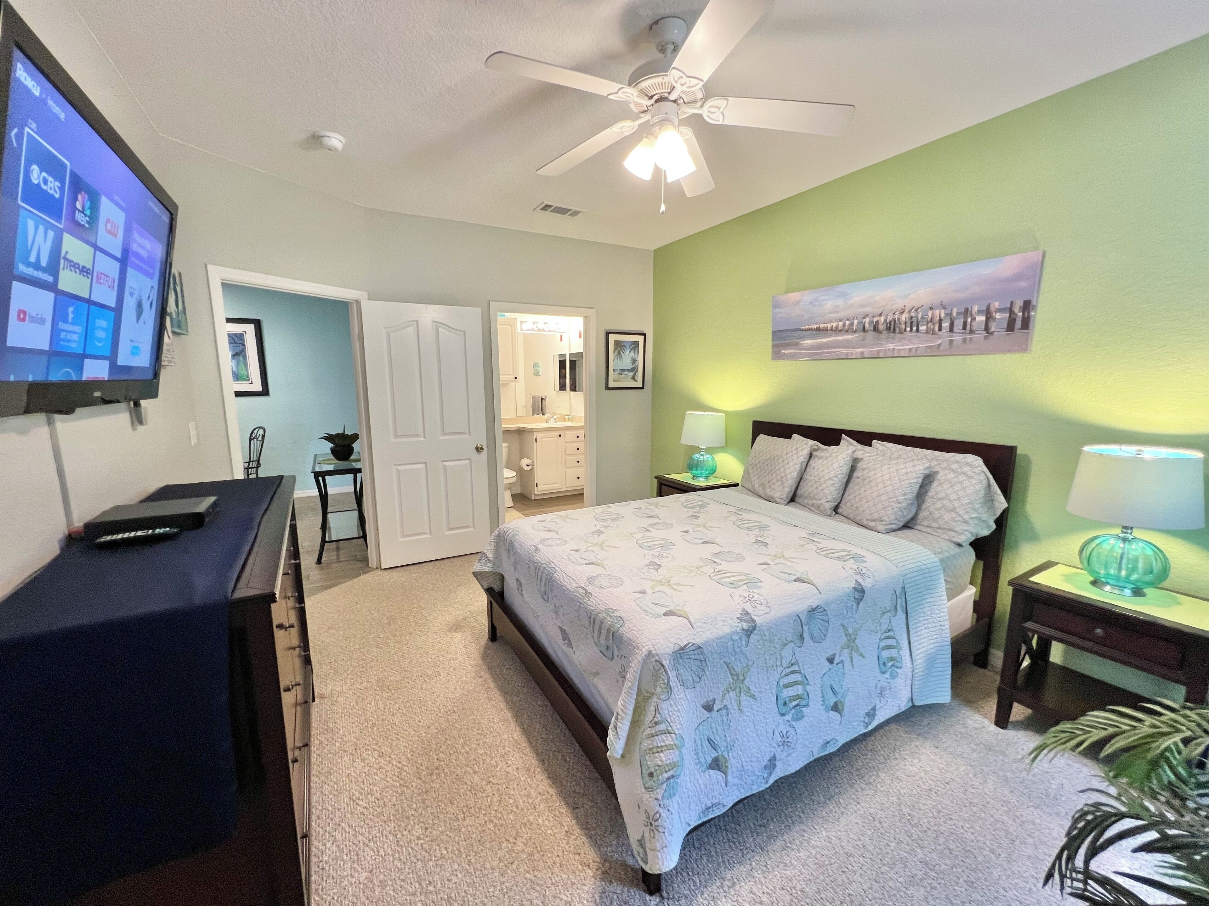 Un dormitorio con cama, escritorio, televisor, ventilador de techo y baño visible a través de una puerta abierta.