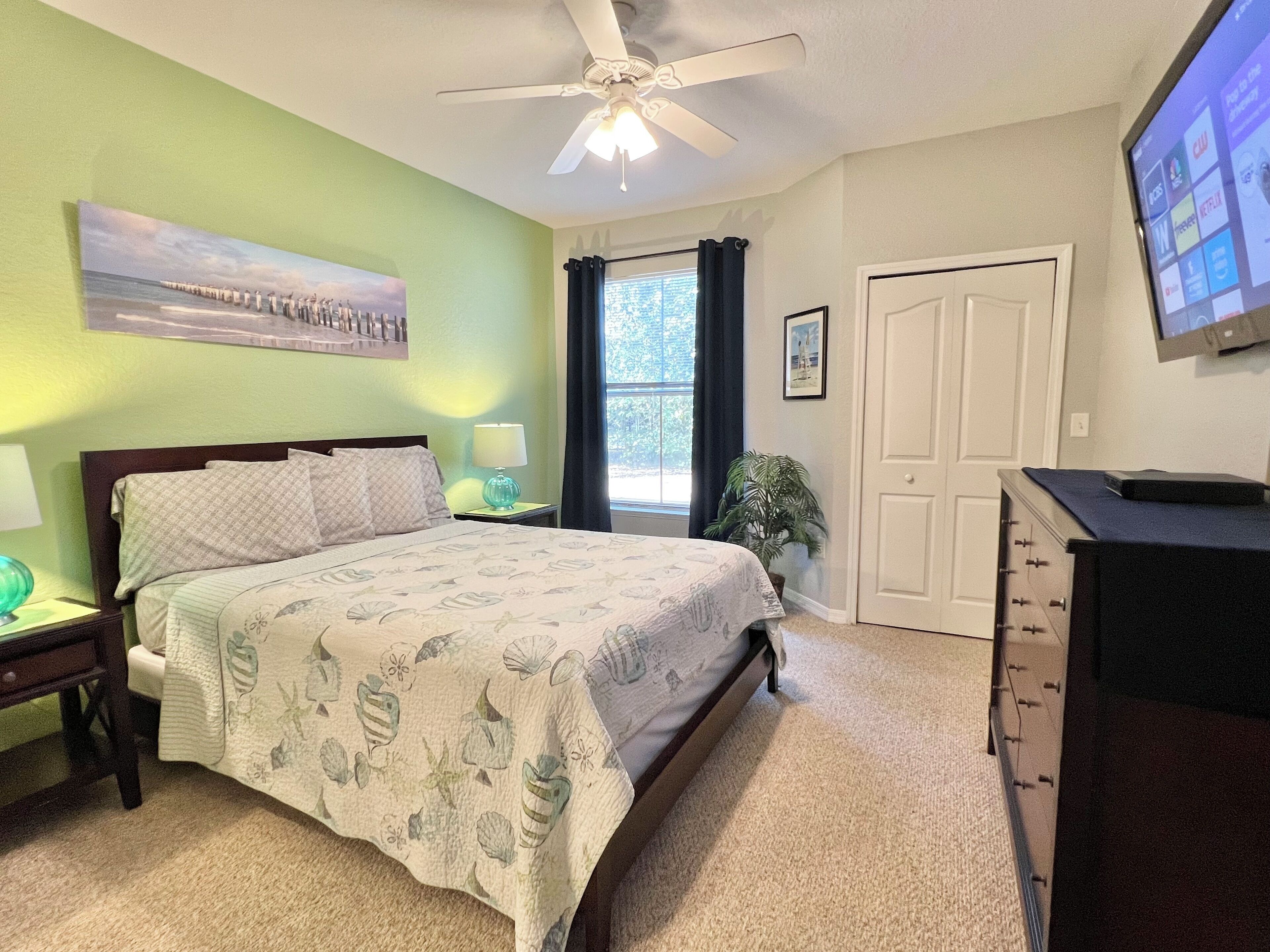 A bedroom with a bed, a nightstand, a lamp, a ceiling fan, a window with curtains, a door, and a TV on the wall.