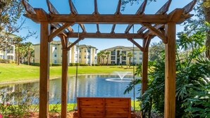 A wooden swing set under a pergola with a view of a pond and buildings in the background.