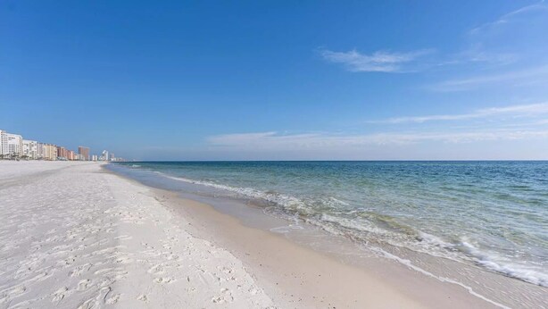 A wide sandy beach with gentle waves, a clear blue sky, and a row of buildings in the background. -  Gulf Shores - AFFORDABLE BEACH VACATION (Gulf Shores)