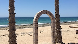 Una estructura en forma de arco con azulejos en una playa arenosa, con palmeras y el océano al fondo.