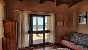 Una habitación con un gran ventanal corredizo que da a vista de playa, un armario de madera, un sofá colorido y un cuadro en la pared.