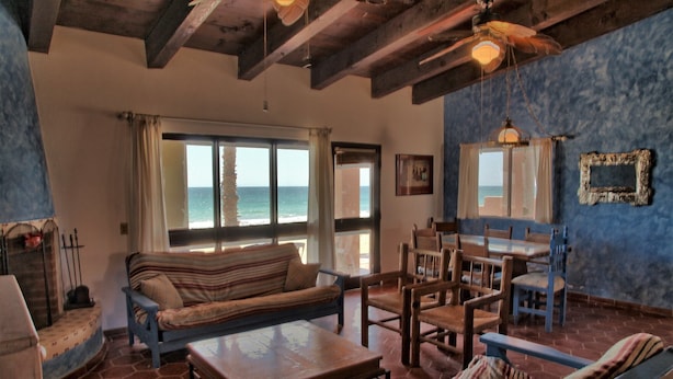 Una sala de estar acogedora con un sofá, una mesa de madera y un comedor con sillas. Hay un ventanal con vistas al mar.