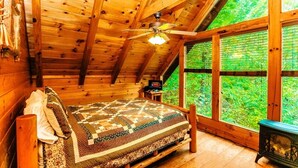 Ein gemütliches Blockhauskabinenzimmer mit Holzbalkendecke, einem Bett mit Patchworkdecke, einem kleinen Tisch und einem Fenster mit Blick auf Grünflächen.