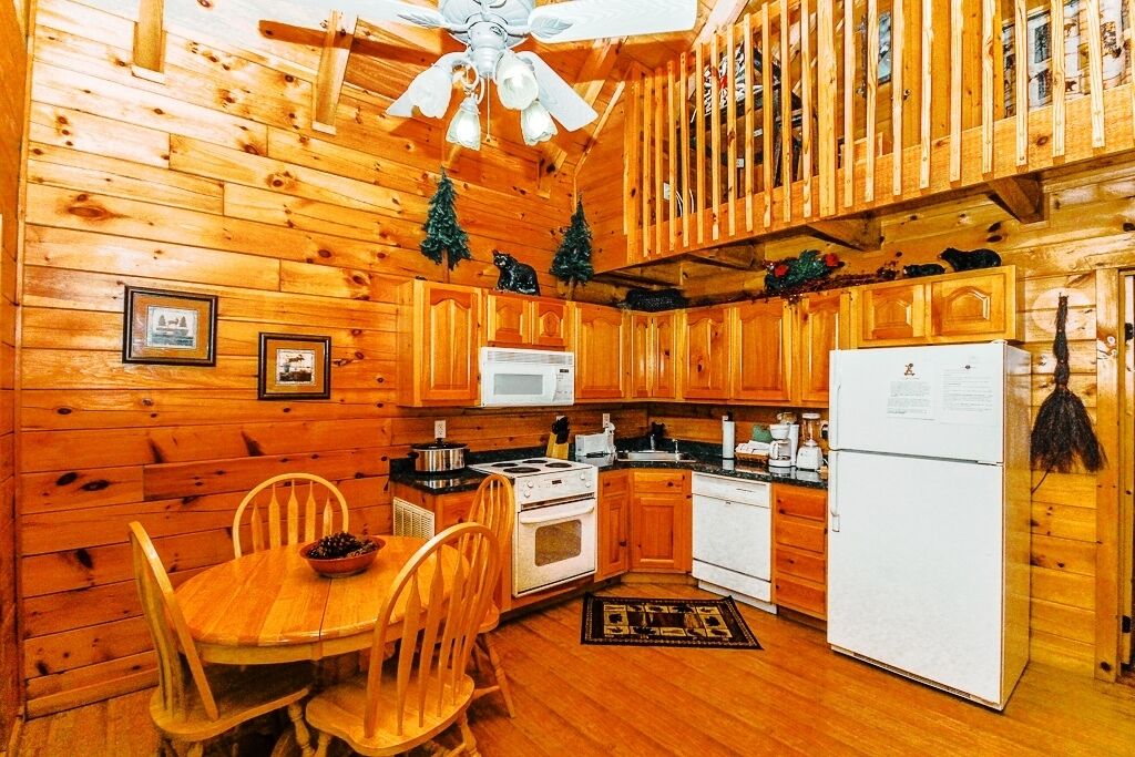 A cabin-style kitchen with wooden walls, a dining table with chairs, a ceiling fan, and a refrigerator.