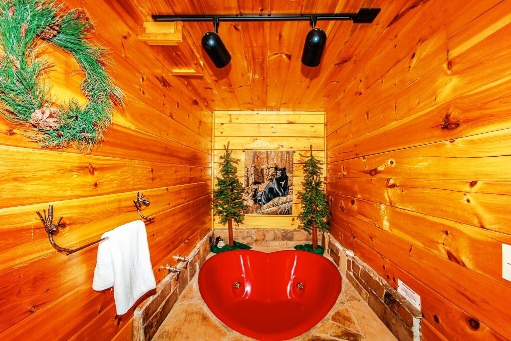 A wooden cabin interior with a heart-shaped bathtub, pine wreath, and two small Christmas trees.