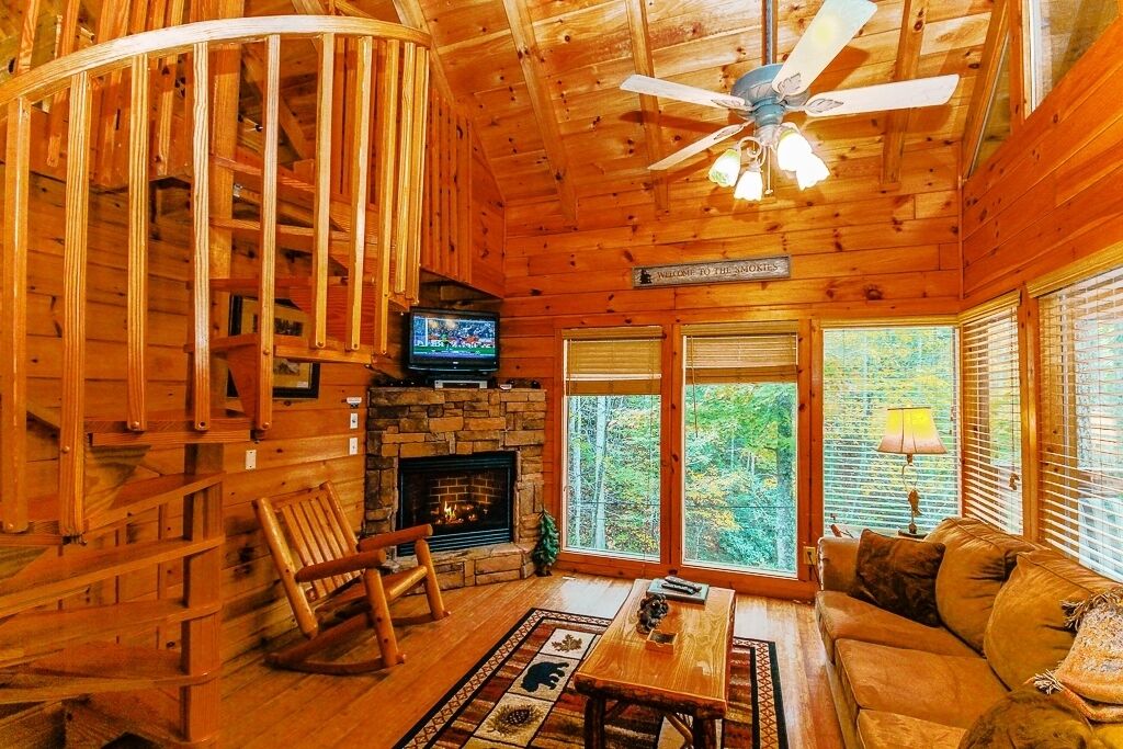 A wooden cabin interior with a stone fireplace, a television, a rocking chair, a sofa, and large windows offering a view of the forest.