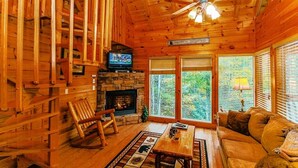Ein Holzblockhaus-Interieur mit Kamin aus Stein, einem Fernseher, einem Schaukelstuhl, einer Couch und großen Fenstern mit Blick auf den Wald.