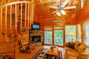 A wooden cabin interior with a stone fireplace, a television, a rocking chair, a sofa, and large windows offering a view of the forest.