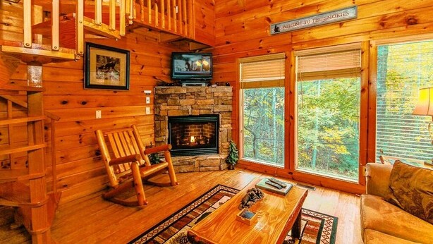 Ein gemütliches Blockhaus-Interieur mit Kamin aus Stein, Fernseher, Schaukelstuhl und großem Fenster mit Blick auf den Wald.