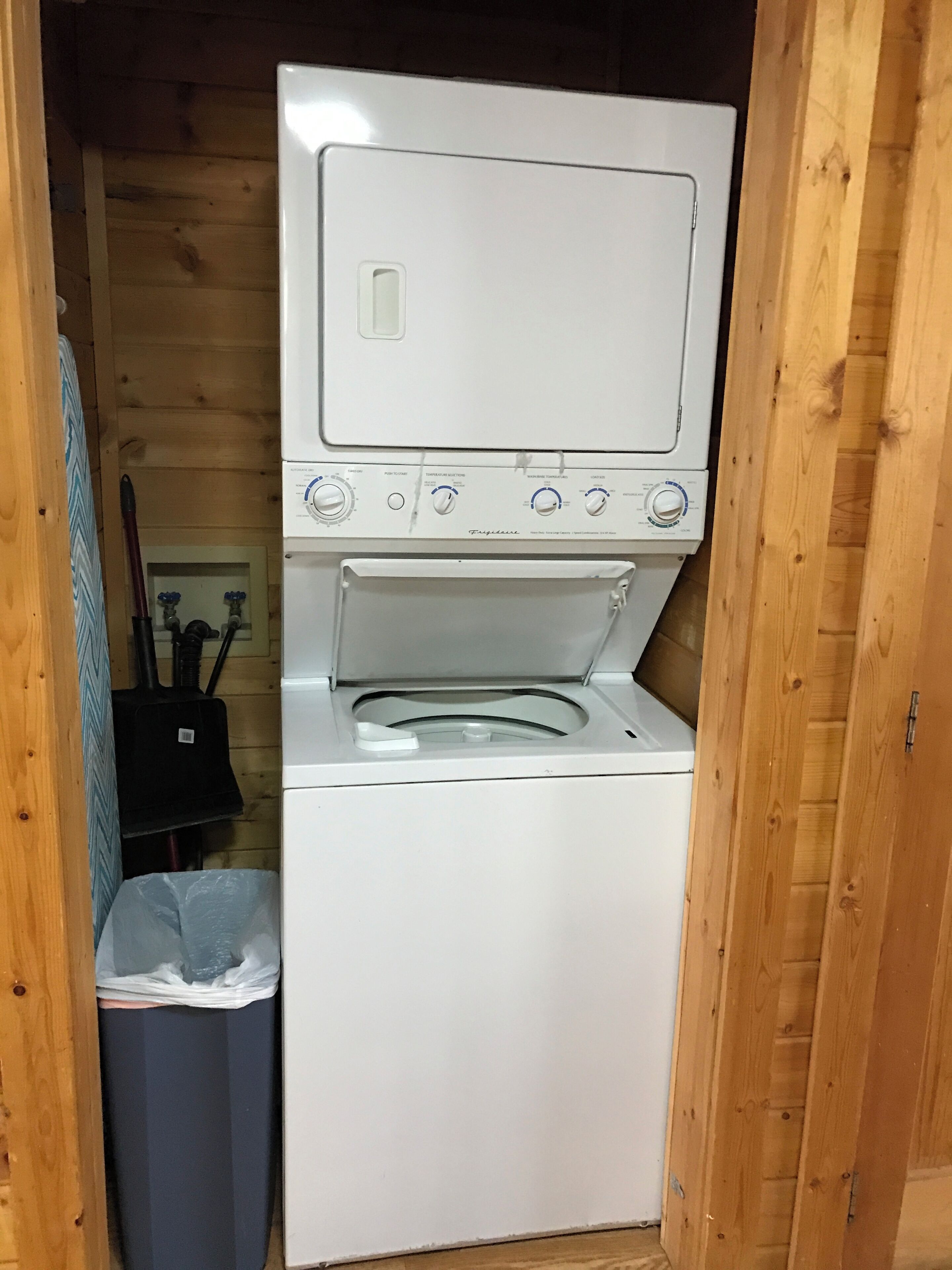 A compact, white laundry room with a built-in washer and dryer, wooden walls, and a trash bin.
