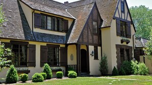 Une maison traditionnelle à colombages, dotée d’un toit à forte pente, de nombreuses fenêtres avec volets et d’un jardin bien entretenu à l’avant, où les arbustes sont taillés avec soin.