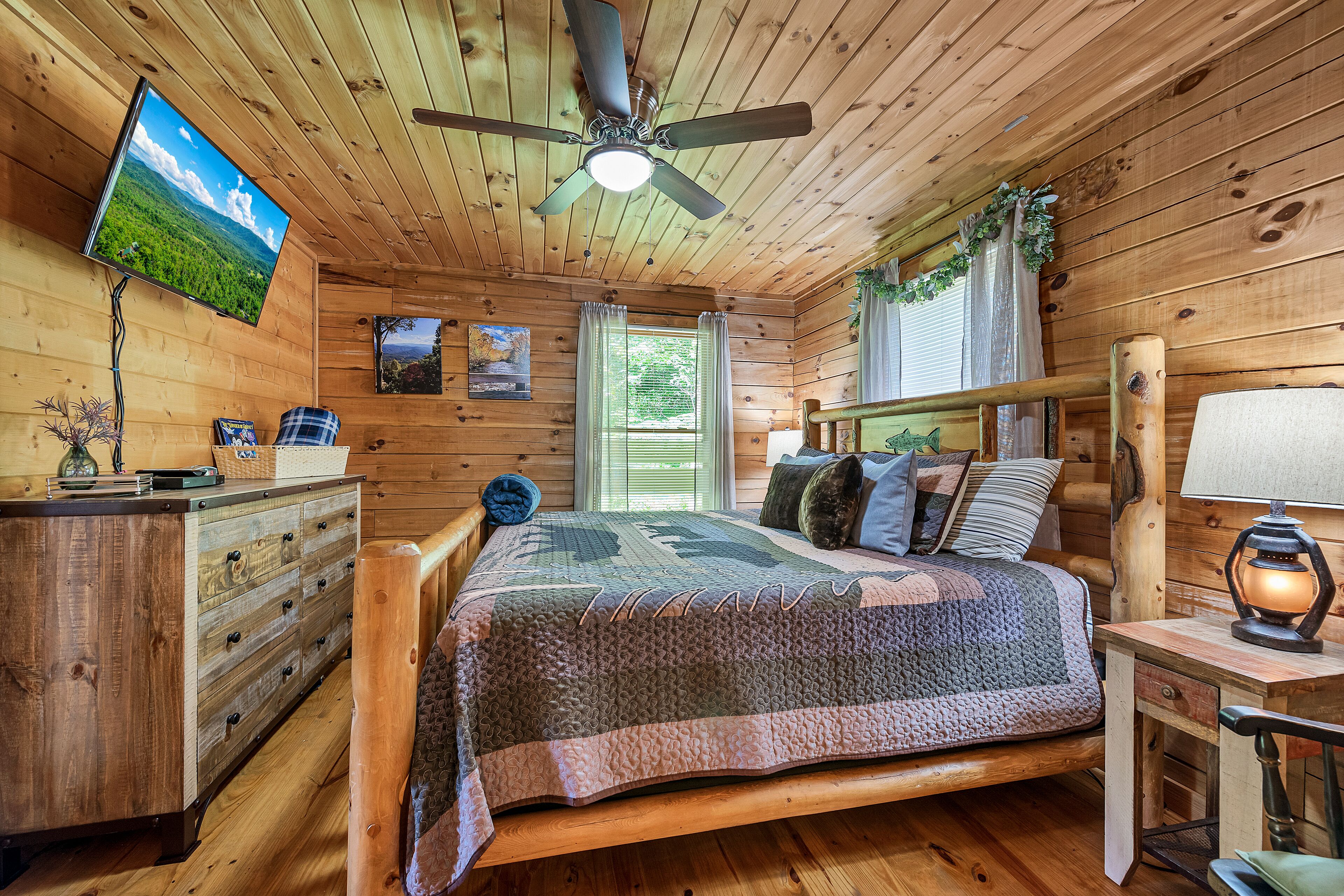 A wooden cabin bedroom with a bed, dresser, TV, and ceiling fan.