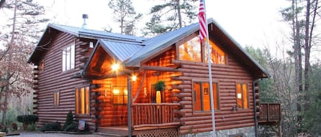 A log cabin with a porch, a stone foundation, and an American flag.