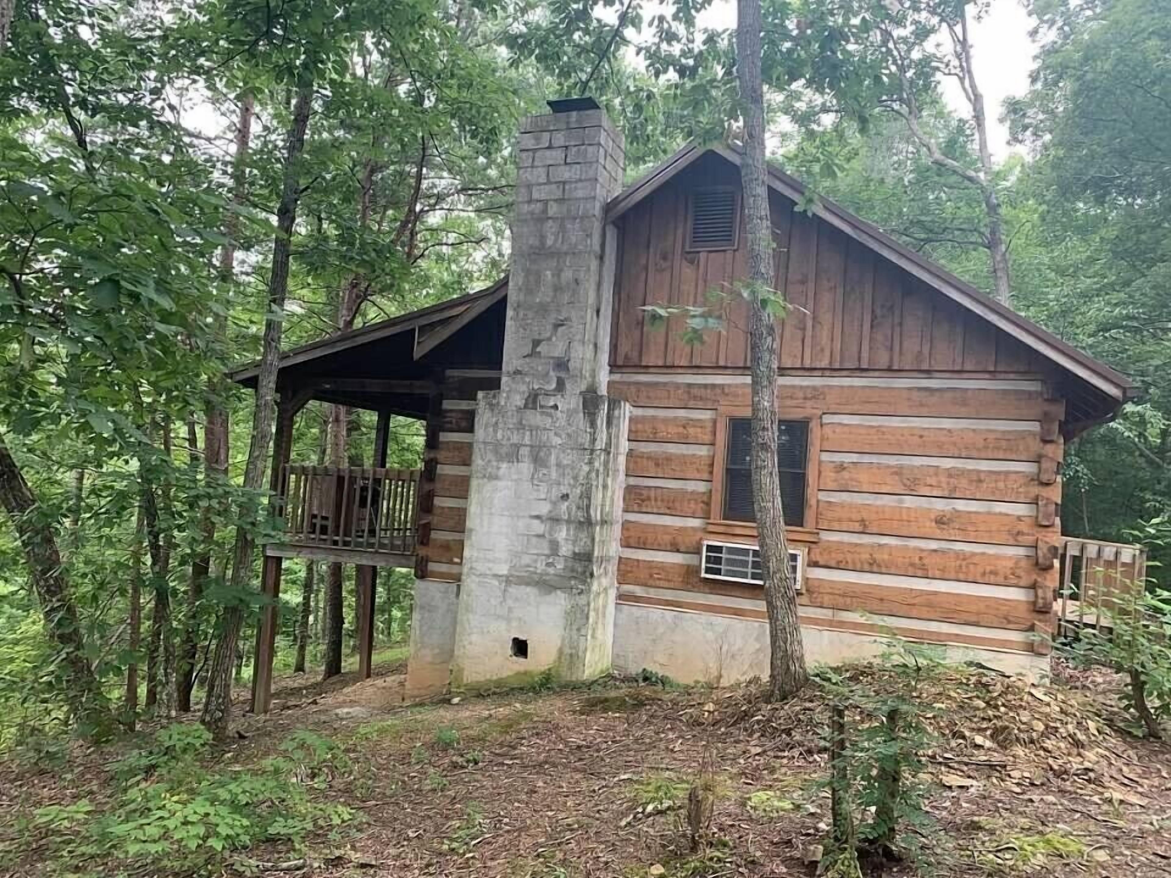 A rustic log cabin with a stone chimney, a wooden deck, and a forested backdrop.