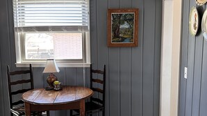 Zona pranzo con tavolo rotondo in legno, due sedie, una finestra con persiane, un quadro e una porta.