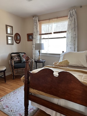 A bedroom with a wooden bed, a wicker chair, a small table with a lamp, and a window with curtains.