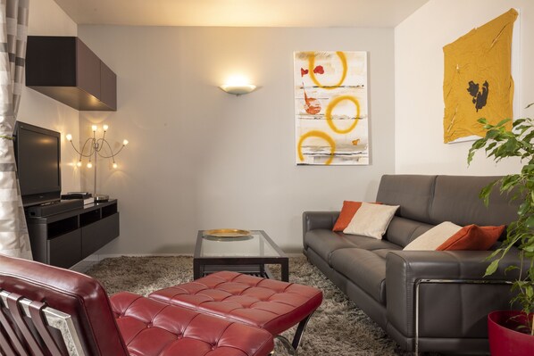 A modern living room with a grey sofa, red leather ottoman, and a glass coffee table. There is a flat-screen TV, a wall-mounted light, and abstract artwork on the wall.