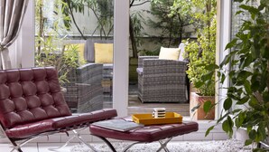 A modern living room with a leather sofa, a wicker chair, and a glass door leading to an outdoor area with plants and a wicker sofa.