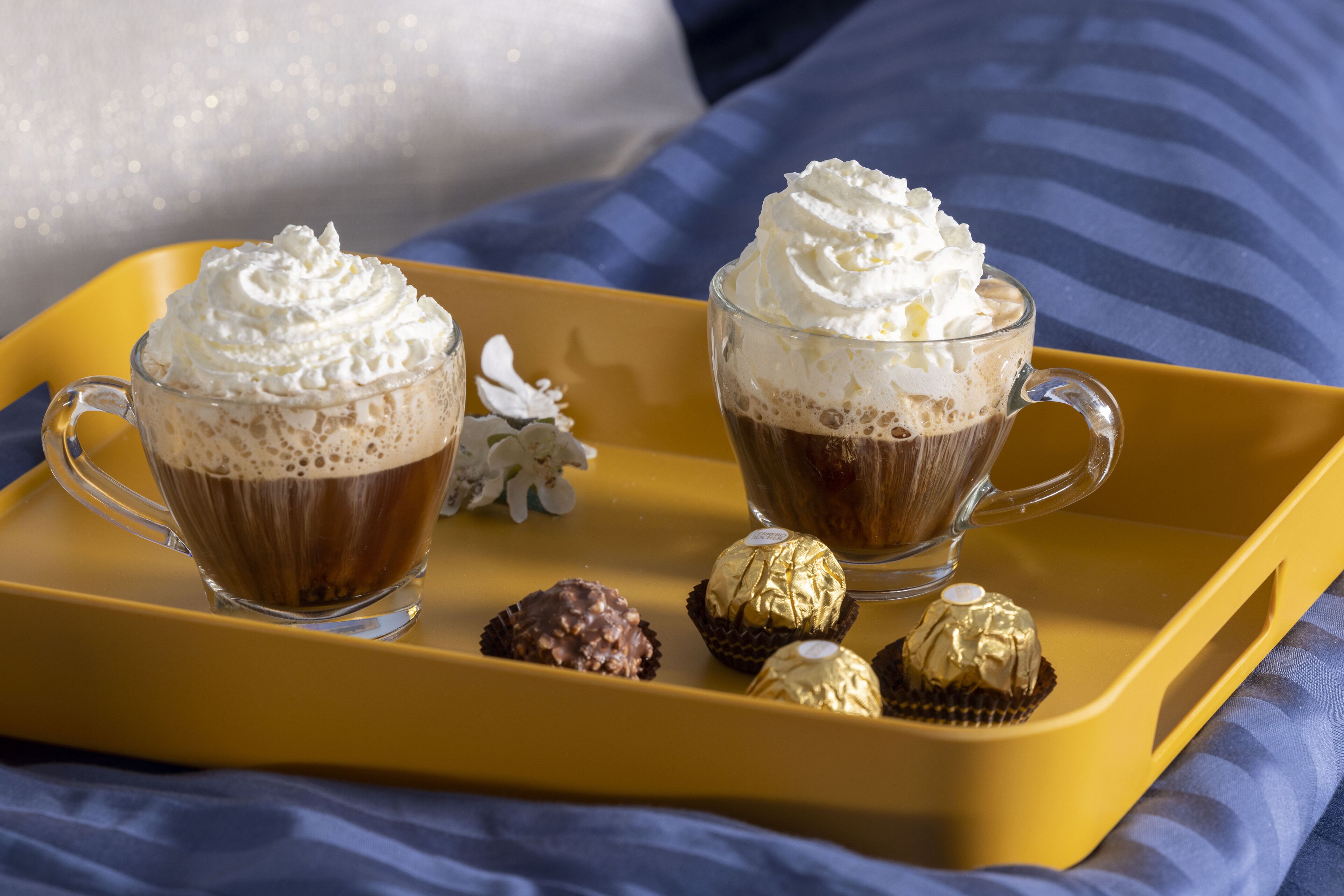 Two cups of coffee with whipped cream on a yellow tray, accompanied by two wrapped chocolates.