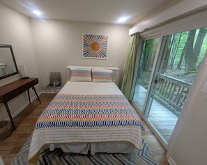 Een slaapkamer met een groot bed, een bureau, een stoel, een raam met uitzicht op bomen en een zonsopgang kunstwerk aan de muur.