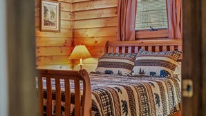 Interior de una cabaña de madera con una cama, una lámpara y una ventana con cortinas.