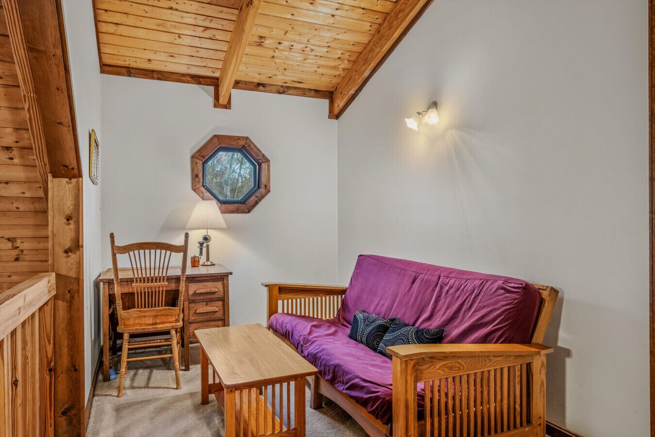 Una habitación acogedora con techo de madera, un sofá morado, una silla de madera, una mesita de madera y una lámpara fijada a la pared.