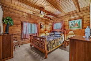 Une chambre dans un chalet en rondins, avec un lit en bois, deux lampes de chevet, un ventilateur de plafond et un tableau encadré au mur.