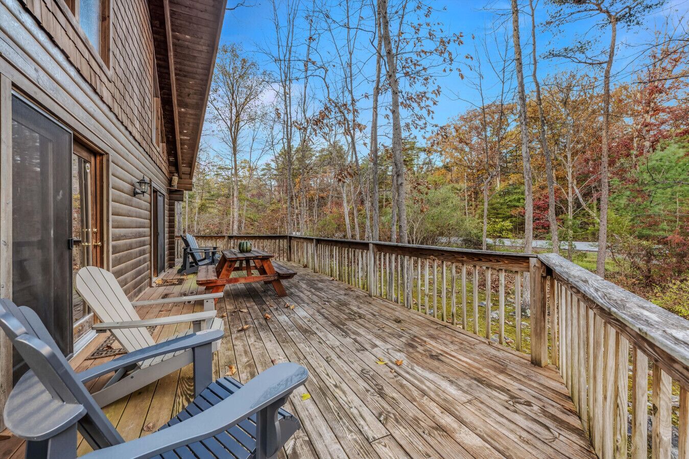 A wooden deck with Adirondack chairs and a picnic table, surrounded by a wooden railing and trees.