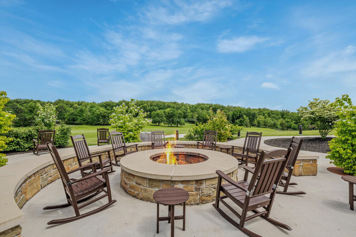 An outdoor seating area with a circular stone fire pit surrounded by wooden chairs and a table.